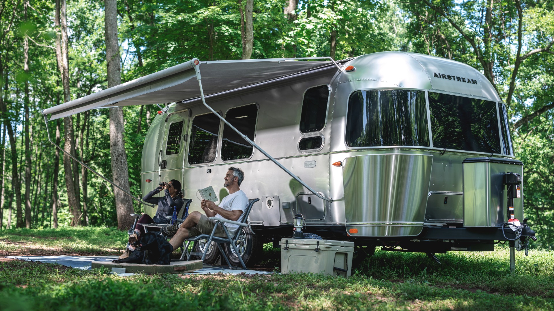 A couple and their dog relaxing beneath the awning of an Airstream Flying Cloud in a wooded campsite